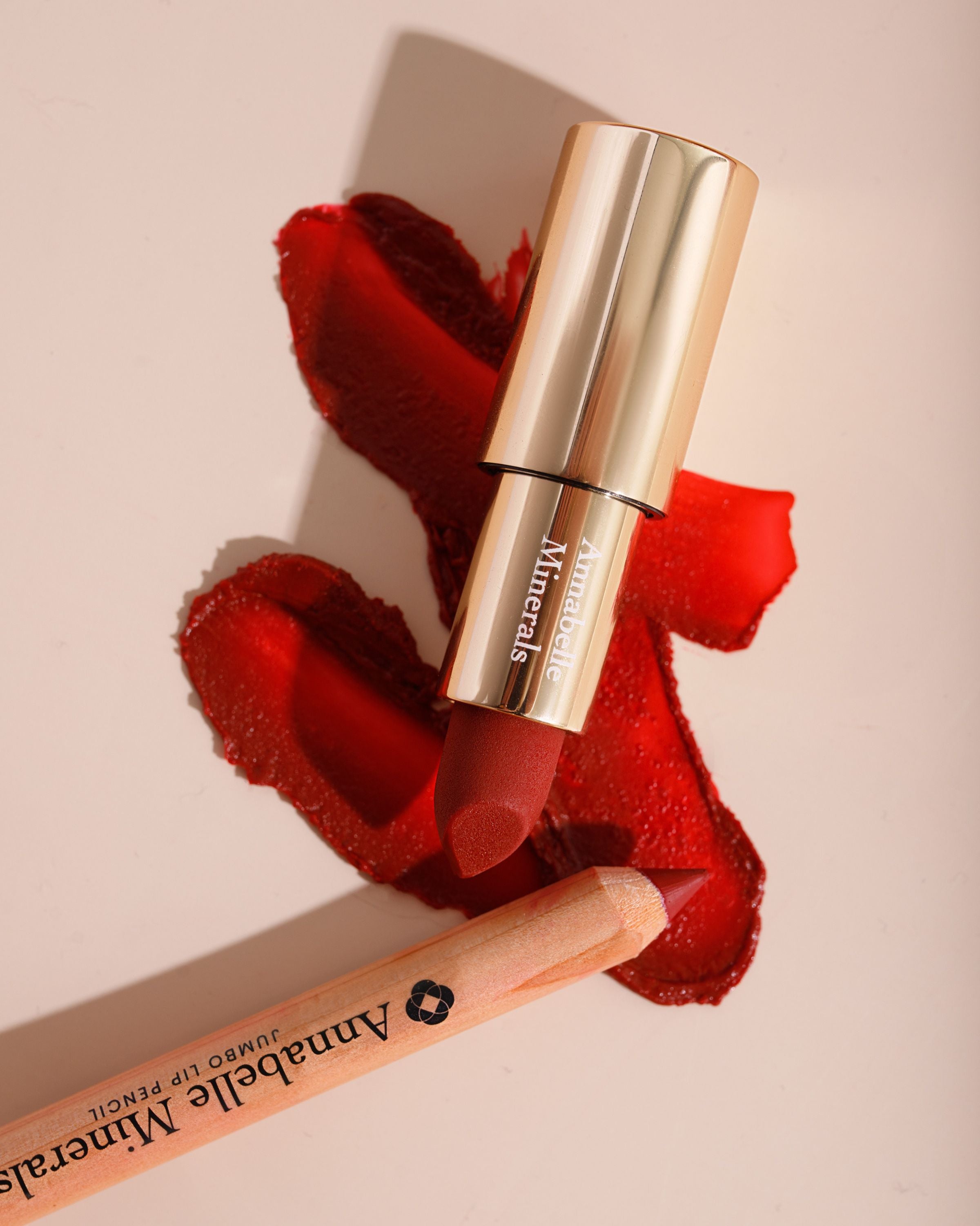 Kosmetyki do ust w czerwonych odcieniach: pomadka Strawberry i kredka do ust jumbo Dahlia Annabelle Minerals na beżowym tle