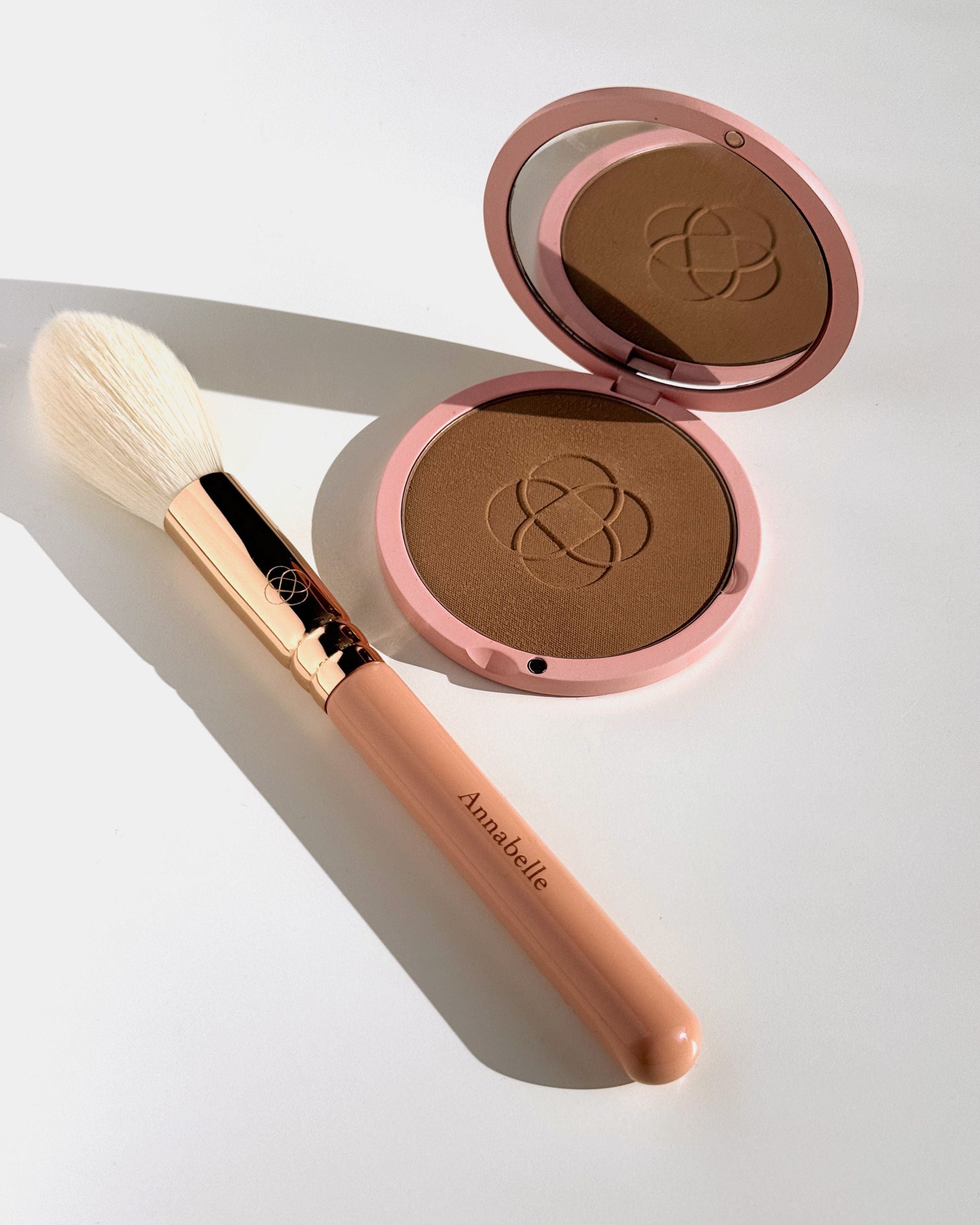 Prasowany bronzer Annabelle Minerals w różowej puderniczce z lusterkiem z pędzlem do rozświetlacza z jasnym włosiem i różową rączką na białym tle