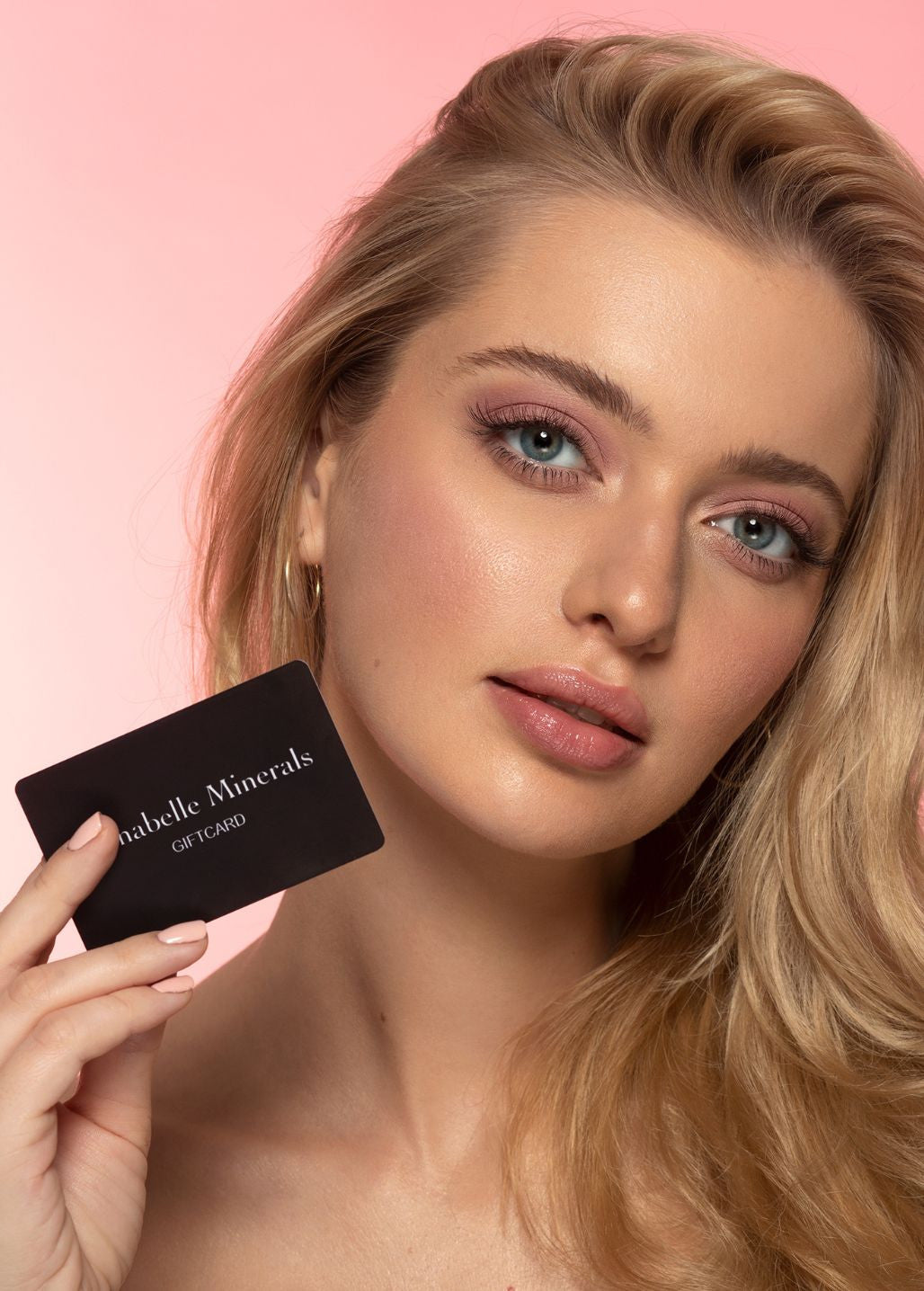 Osoba z długimi, blond włosami trzyma czarną kartę podarunkową Annabelle Minerals na różowym tle