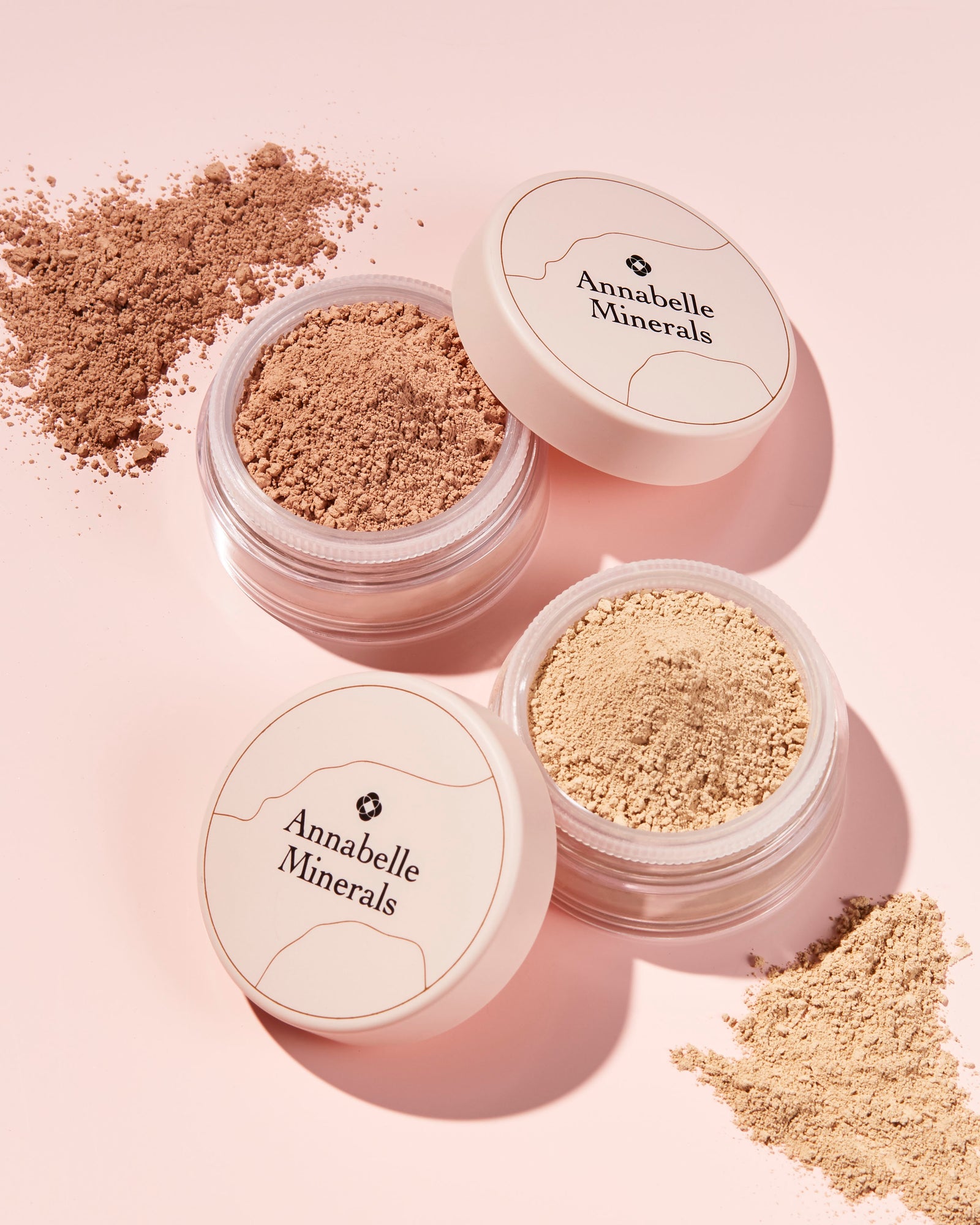Trzy pojemniki z minerałami Annabelle Minerals na jasnoróżowym tle, wokół rozsypany podkład.