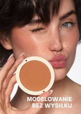 Osoba z figlarną miną trzyma bronzer Annabelle Minerals w odcieniu Toasted, z napisem MODELOWANIE BEZ WYSIŁKU